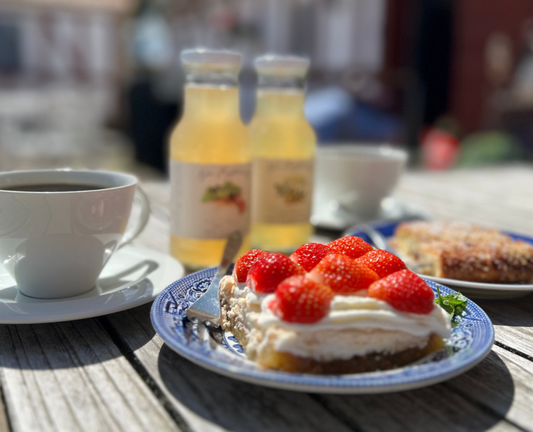A cup of coffee with accompanying cake on a table in the sunshine, with juice from Nybro Fruit Plantation visible in the background.